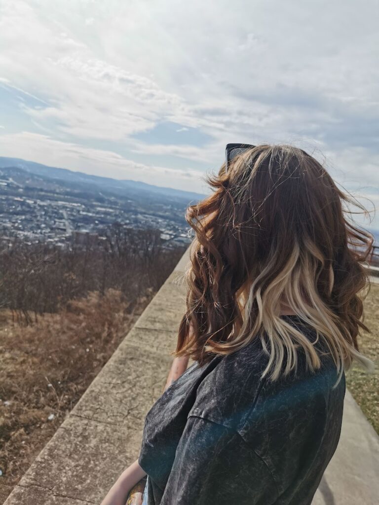 Alyssa admiring a stunning cityscape from a hilltop, back facing the camera. Wind tousles her hair as she takes in the breathtaking view.